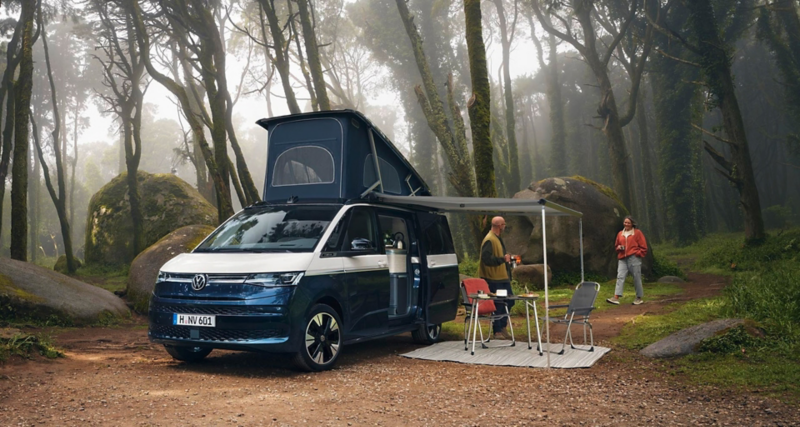 Ein VW California parkt in einem Wald. Die Markise ist aufgestellt, darunter steht eine Campinggarnitur.