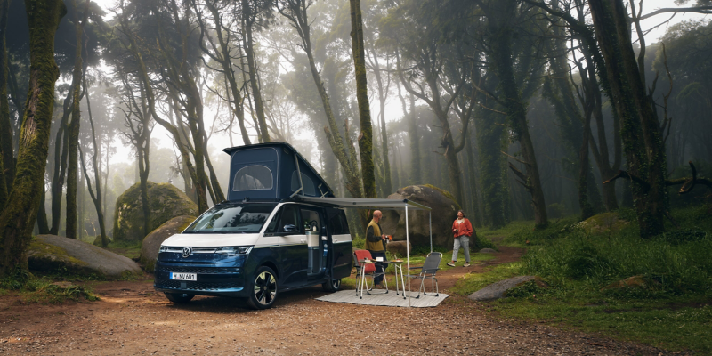 Ein VW California mit aufgestelltem Hochdach und ausgefahrener Markise steht in einem Wald und zwei Menschen campen.