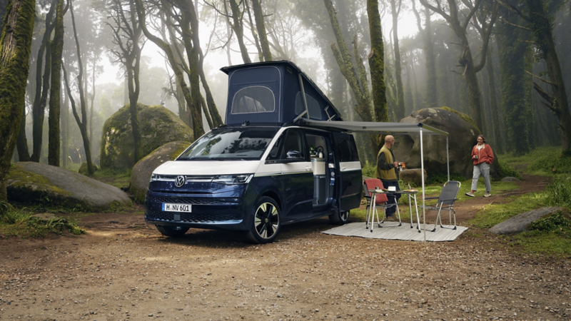 Ein VW California Parkt mit aufgebauter Markise in einem Wald.