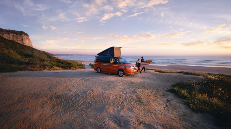 Ein VW California steht mit aufgestelltem Dachbett am Strand. Im Hinterfgrund sehen wir zwei Surfer.