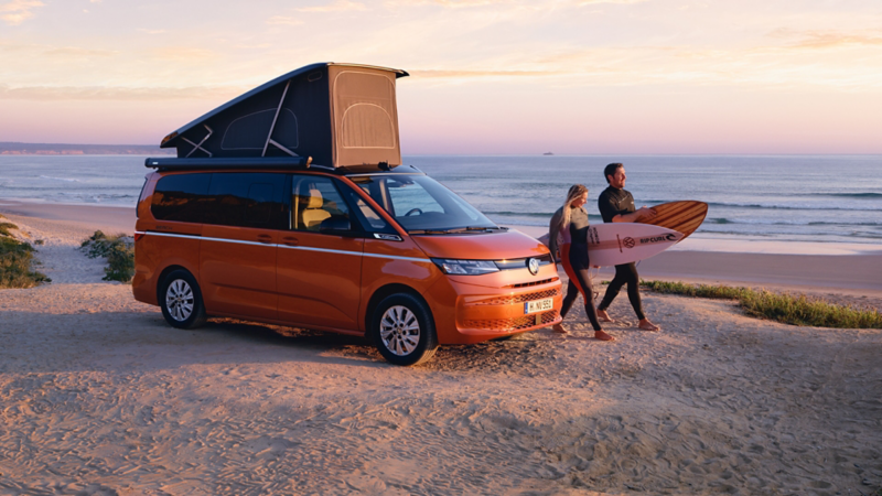 VW California steht mit aufgestelltem Dachzelt an einem Strand. Daneben gehen zwei Surfer.