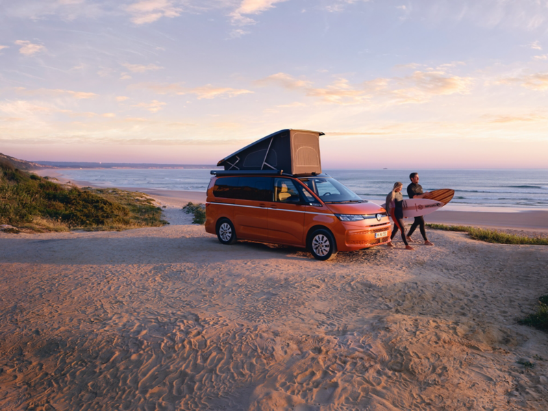 Ein VW California Beach steht am Strand und Surfer machen sich auf den Weg.