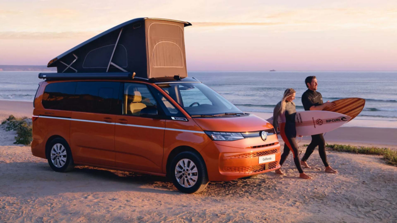 Couple walking with surfboards beside a Volkswagen California camper van parked on a beach at sunset
