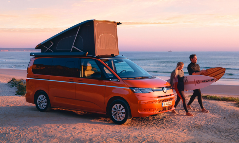 Ein VW California parkt am Strand und ein Pärchen mit Surfbrettern geht vorbei.