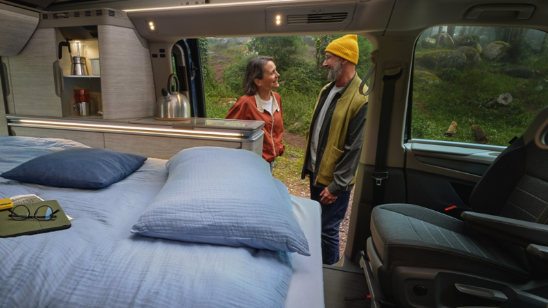 Man and woman standing in front of their VW California in a wood. The bed in the rear is made up and the sliding door is open.