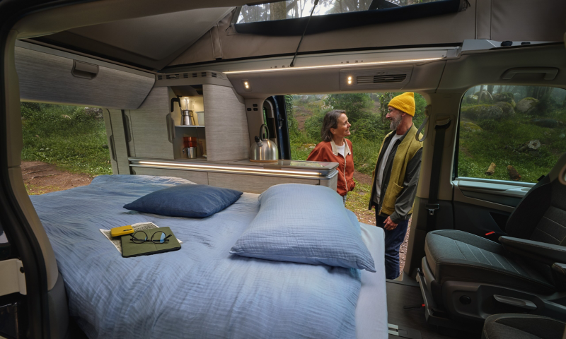 Une femme et un homme se tiennent à côté d’une VW California dans laquelle un lit a été installé.