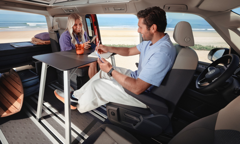 Een man en een vrouw zitten aan het klaptafeltje in het interieur van een VW California.