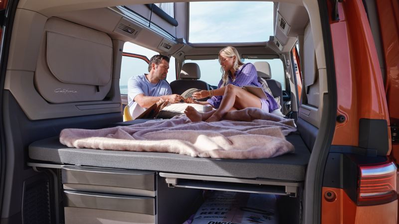 Een vrouw en een man zitten op het uitgeklapte bed van de VW California.