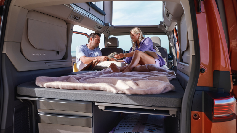 Eine Frau und ein Mann sitzen auf dem aufgebauten Bett des VW California.