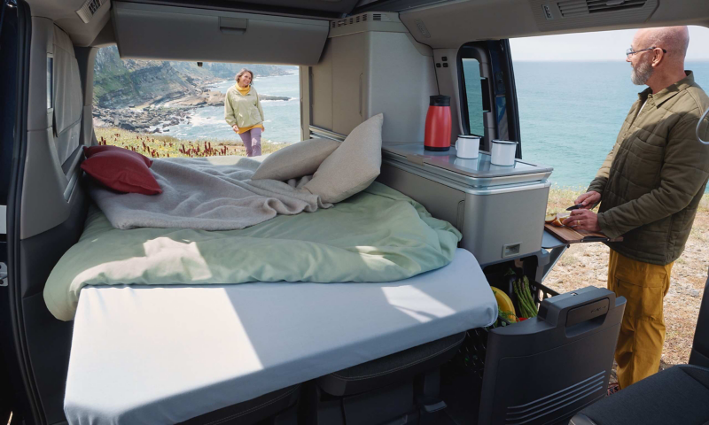 Photo of a California parked on a beach with two people sat in front of it eating.