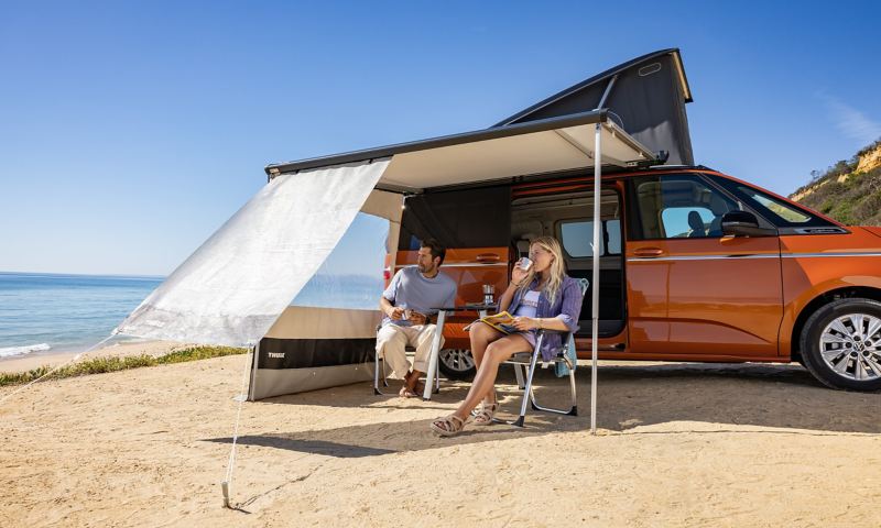 Ein Mann und eine Frau sitzen auf Campingstühlen unter der Markise neben dem VW California.