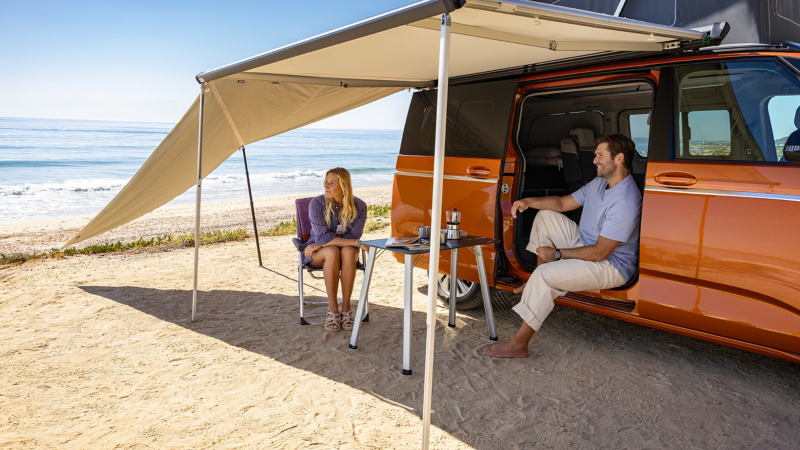 Eine Frau und ein Mann sitzen unter einer aufgebauten Markise des VW California.