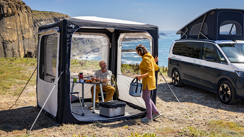 Ein Ehepaar an einem Campingtisch im Dometic Vorzelt neben einem VW California mit aufgestelltem Dachzelt.