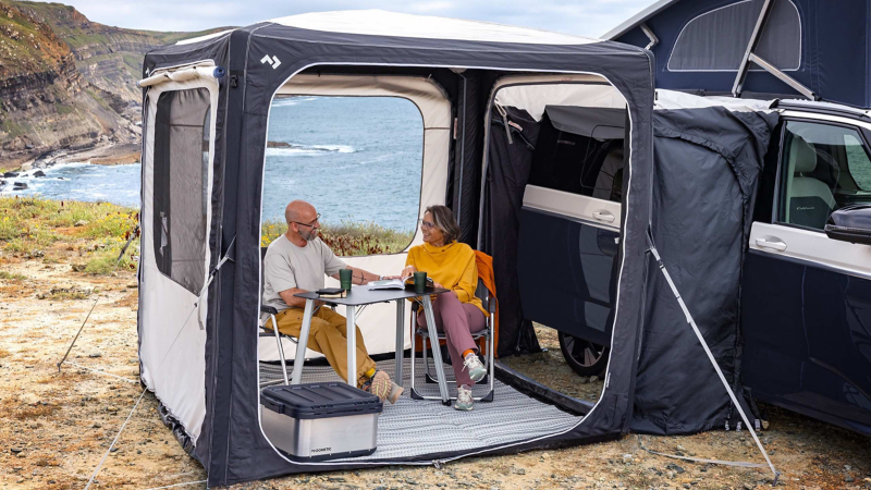 Photo of a California parked on a beach with two people sat in front of it eating.