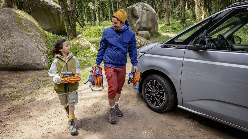 Ein Mann und Kind tragen Stromkabel um den geparkten VW California anzuschließen.