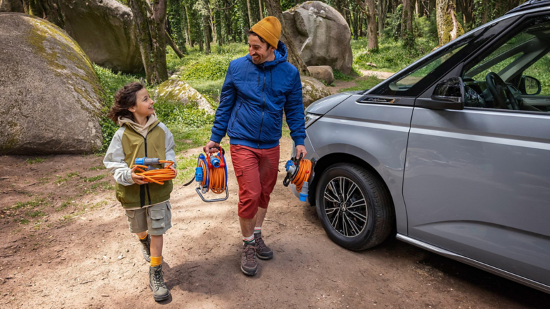 Erwachsener und Kind tragen Campingzubehör neben einem Volkswagen Fahrzeug