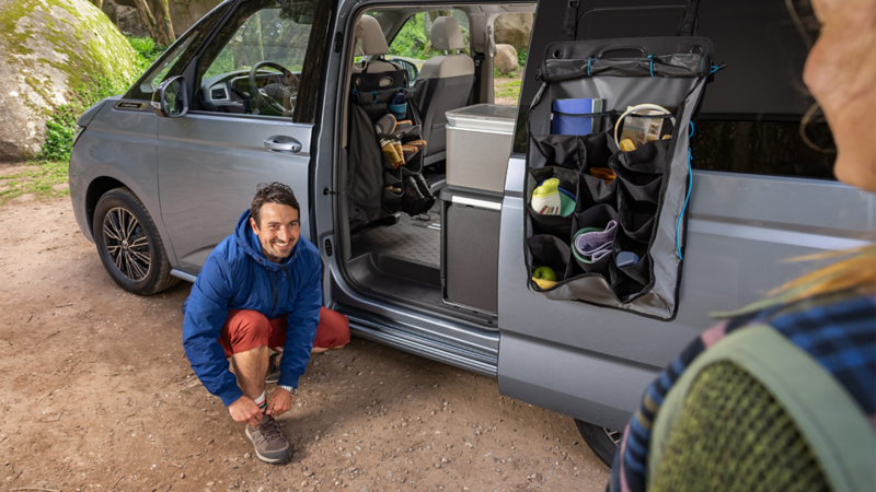 Campingsituation mit VW California in der Natur. Ein Mann bindet sich die Schuhe zu.