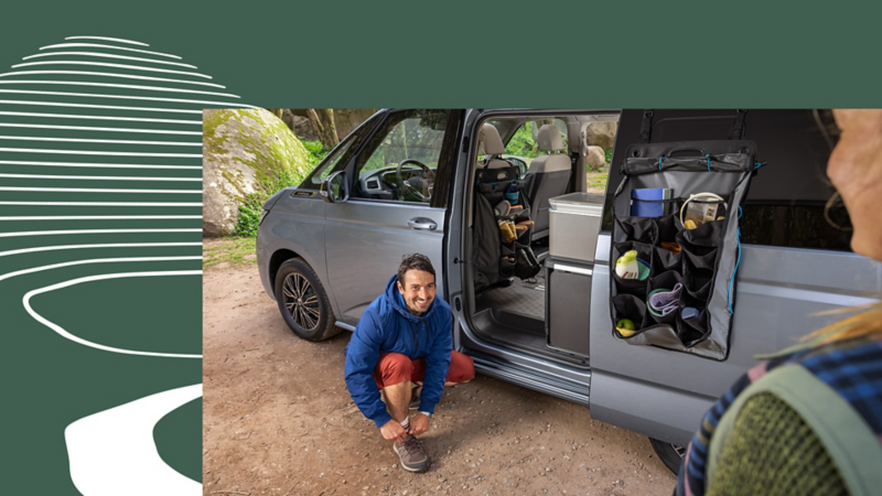 Ein Mann bindet seine Schuhe neben dem VW California COAST. In der geöffneten Schiebetür hängt eine praktische Tasche.