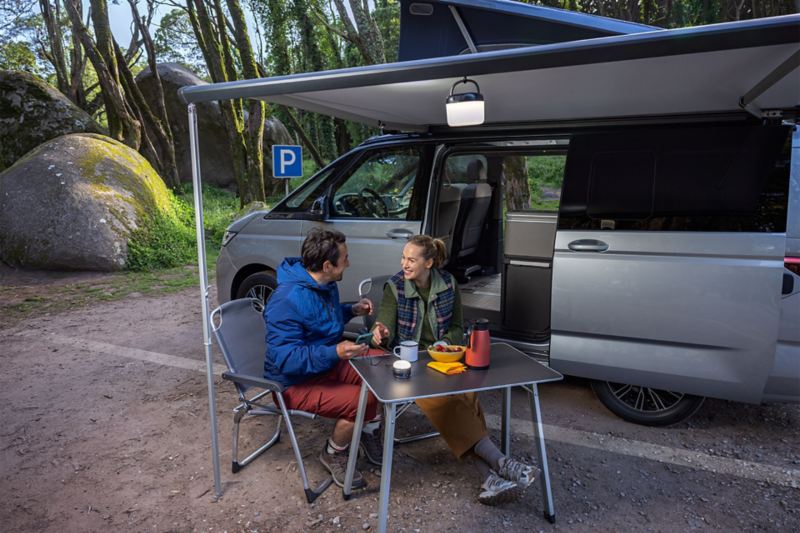 Un homme et une femme assis sur des chaises de camping sous l’auvent à côté du Volkswagen California en forêt.