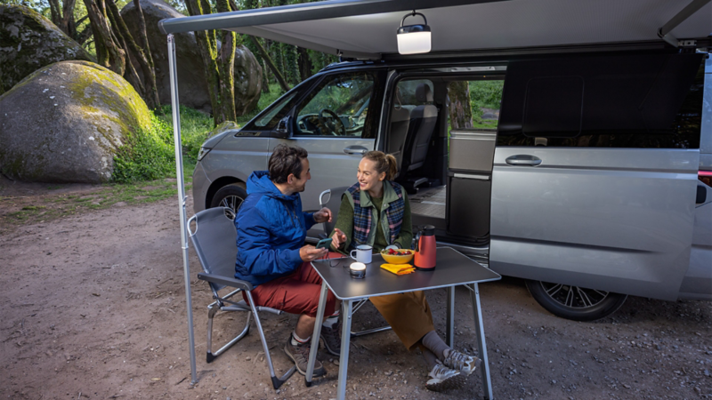 Auf einem Waldcampingplatz sitzt ein Paar am Tisch vor einen VW California. Die Markise ist ausgefahren.