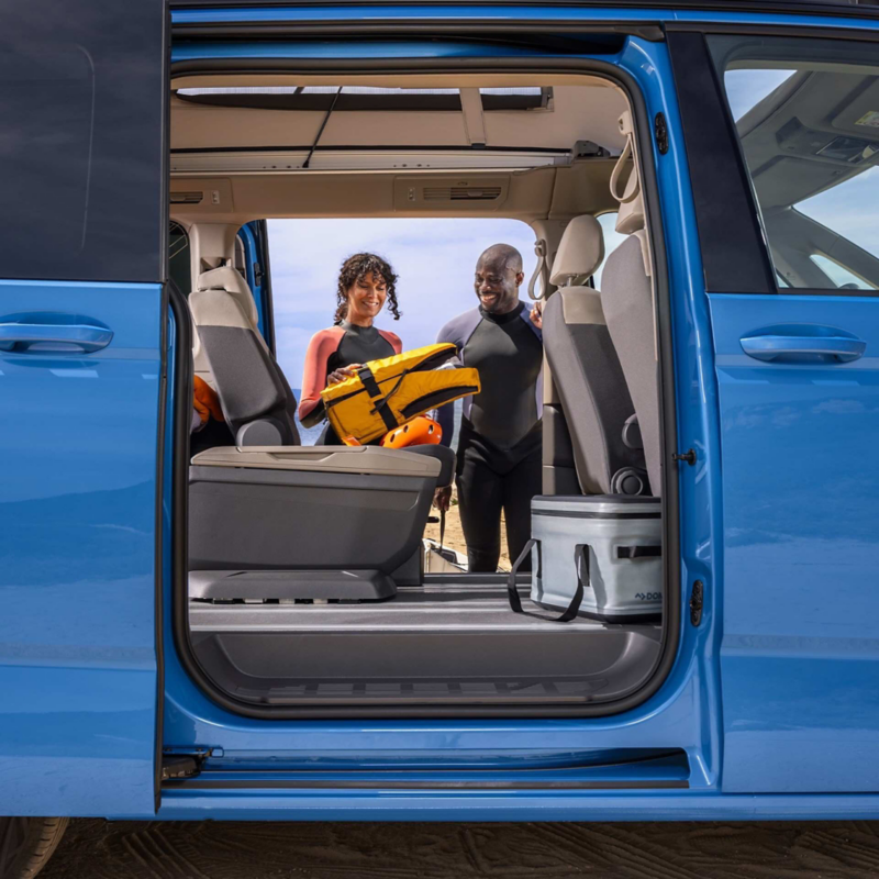 Photo showing a man and a woman wearing swimming gear viewed through the open side doors of a VW California. 
