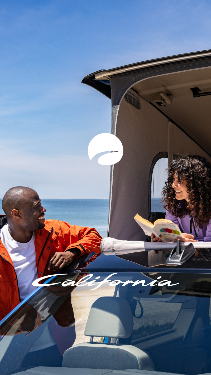 Eine Frau liegt im aufgestellten Dachzelt eines VW Californias und hält ein Buch in der Hand. Ein Mann lehnt in der geöffneten Tür des Fahrzeugs, beide lachen sich an.