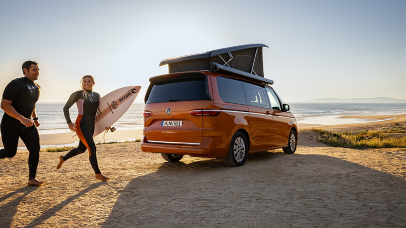 Ein VW California steht am Strand.  Am linken Rand des Bildes kommen zwei Surfer in Neoprenanzügen angerannt. 