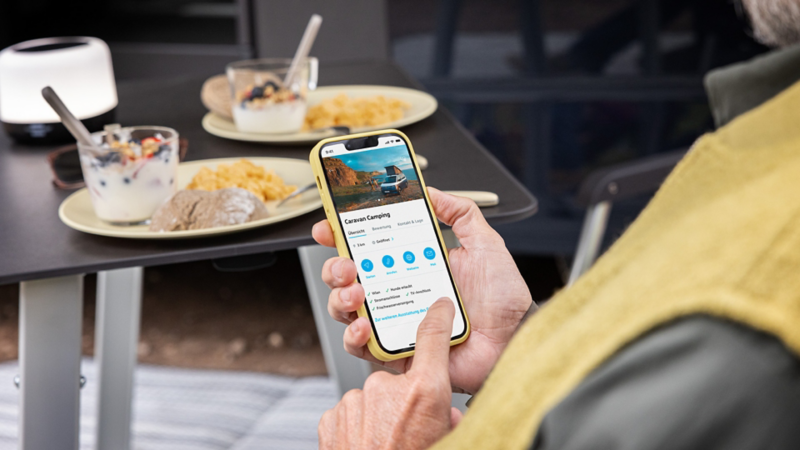 Die Cali on Tour App ist im Close-Up zu sehen. Ein Mann bedient sie mit Smartphone am Campingtisch.
