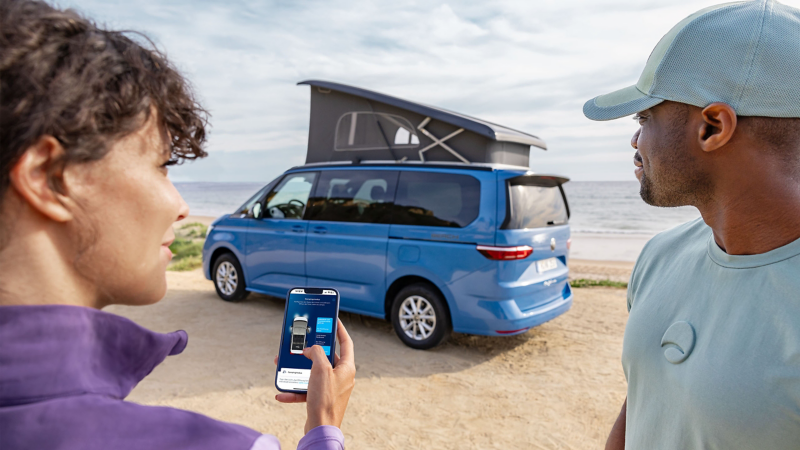 Ein Mann steht neben einer Frau am Strand, während sie das Aufstelldach des VW California per Smartphone fernbedient.