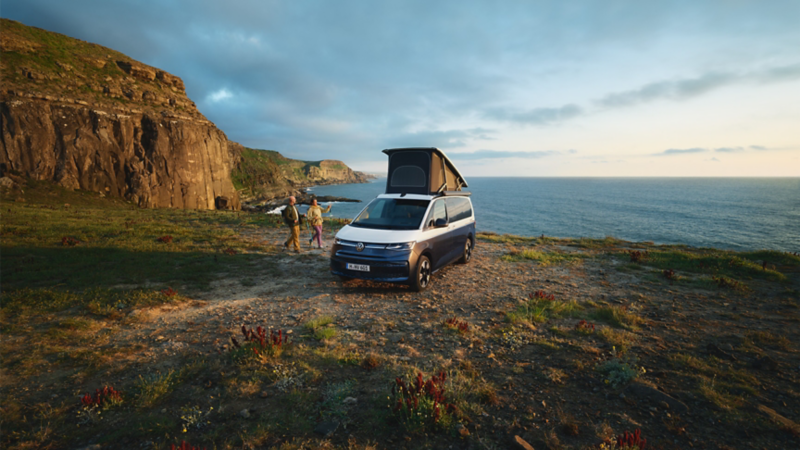 Ein parkender VW California steht mit ausgefahrener Markise und aufgestelltem Dachbett auf einem Plateu an der Steilküste. 