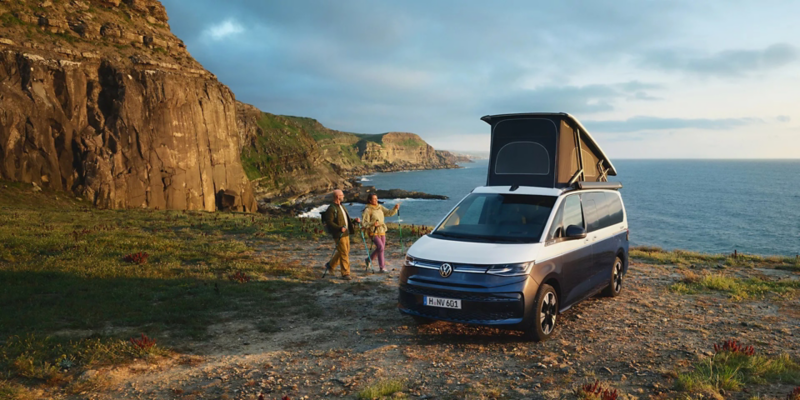 Persone accanto a un Volkswagen California parcheggiato con il tetto sollevato, scogliere e mare sullo sfondo.