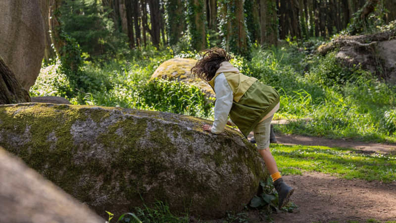 Ein Kind in funktionaler Kleidung klettert im Campingurlaub auf einen großen Stein im Wald.