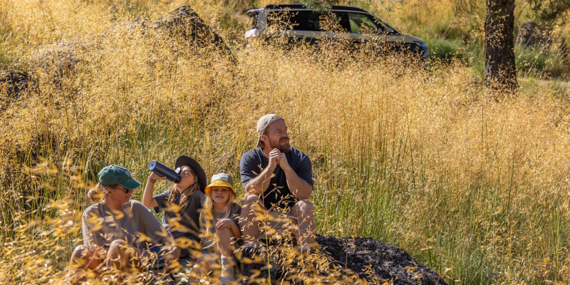 Ein Mann sitzt mit seinen Kindern in der Natur, im Hintergrund parkt ein VW California