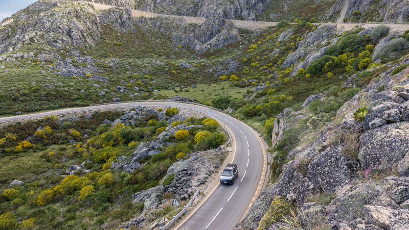 A VW California drives leisurely down a road through a picturesque mountain landscape.