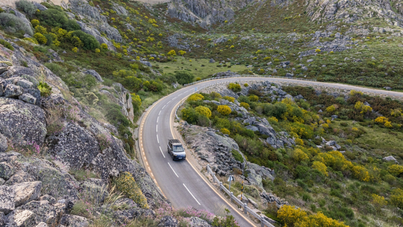 Un véhicule VW California circule sur une route de campagne sinueuse à travers un paysage montagneux.