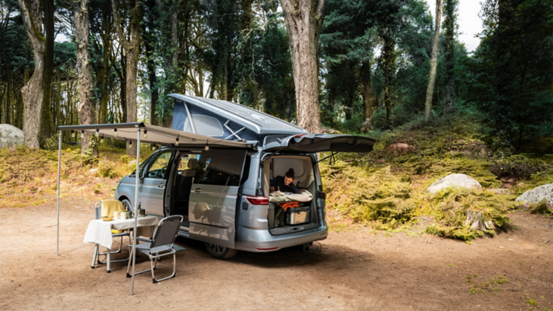 Ein geparkter VW California auf einem Waldcampingplatz, mit ausgefahrener Markise und aufgestelltem Dachbett. Schiebetür und Heckklappe sind geöffnet und auf der Liegefläche liegt eine Frau und liest ein Buch.