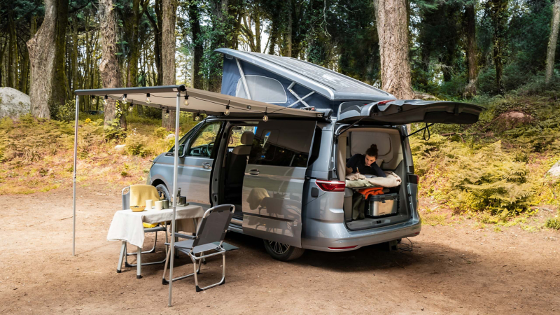 Eine Frau liegt bei geöffneter Heckklappe auf dem Bett des VW California, der Campingbereich ist aufgebaut.