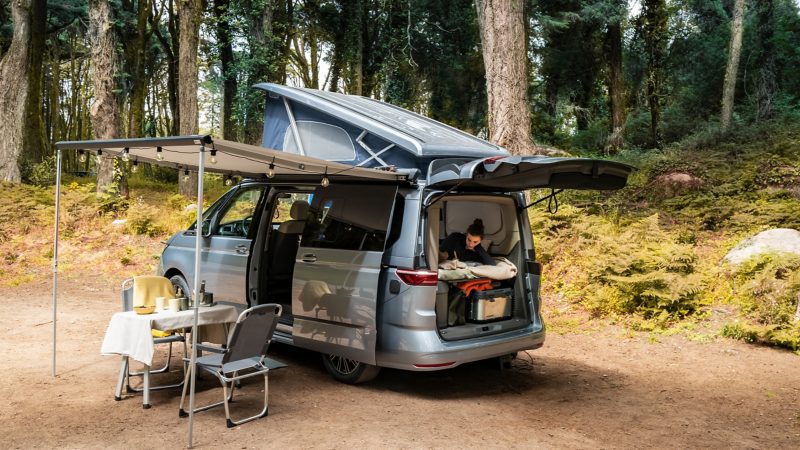 Ein VW California Parkt mit aufgebauter Markise in einem Wald.