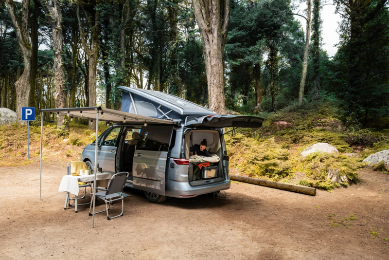 Un Volkswagen California est stationné dans un bois, le toit-tente ouvert, le coffre ouvert et l'auvent déployé. À l'intérieur, une femme est confortablement installée sur le canapé.