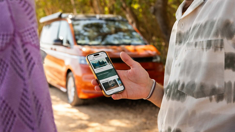 In the foreground, we see two people in close-up. The person on the right is holding a mobile phone with the California app open. In the background, we see a VW California.