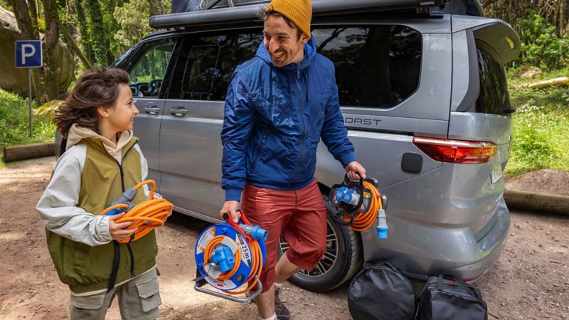 Un homme et un enfant transportant des câbles électriques marchent à côté d'un VW California.