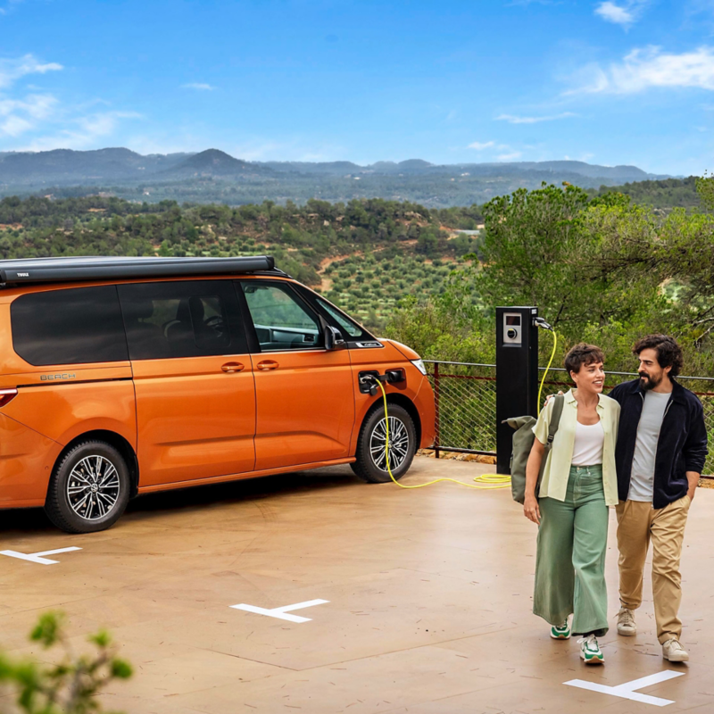 Un couple souriant marche loin d'un Volkswagen Multivan Beach orange garé à une borne de recharge extérieure, avec un paysage montagneux en arrière-plan. Le van est en charge.
