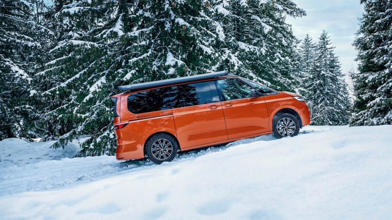 Ein VW California in Energetic Orange fährt durch eine weiße Winterlandschaft, umgeben von verschneiten Nadelbäumen.