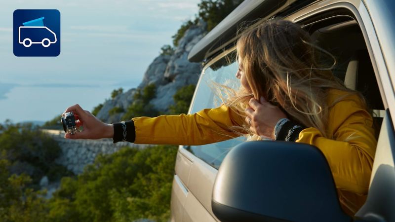 En kvinde fotograferer havet ud ad vinduet på en parkeret VW California 6.1. Ikonet for Cali-on-Tour-appen ses øverst til venstre på billedet.
