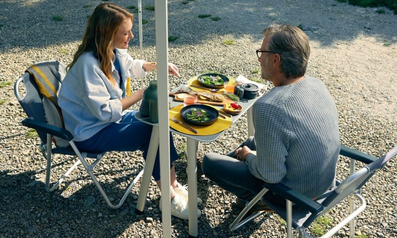 Zwei Personen sitzen auf den Campingmöbeln von VW Grand California und essen etwas.