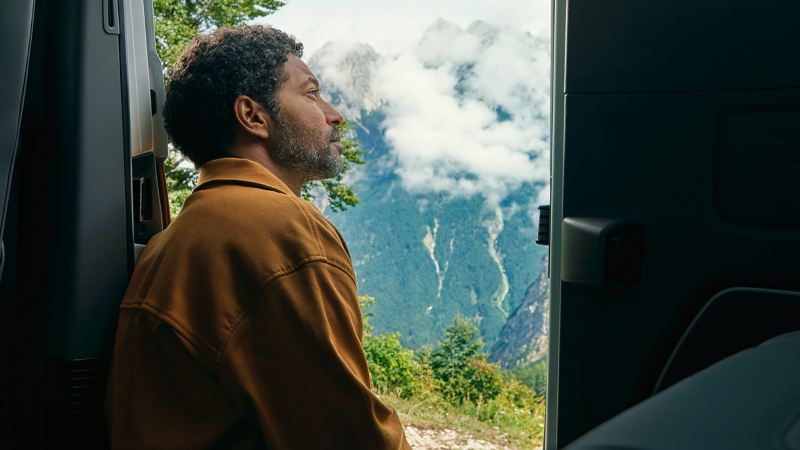 Ein Mann sitzt in der Tür eines California 6.1 und blickt auf eine Berglandschaft.