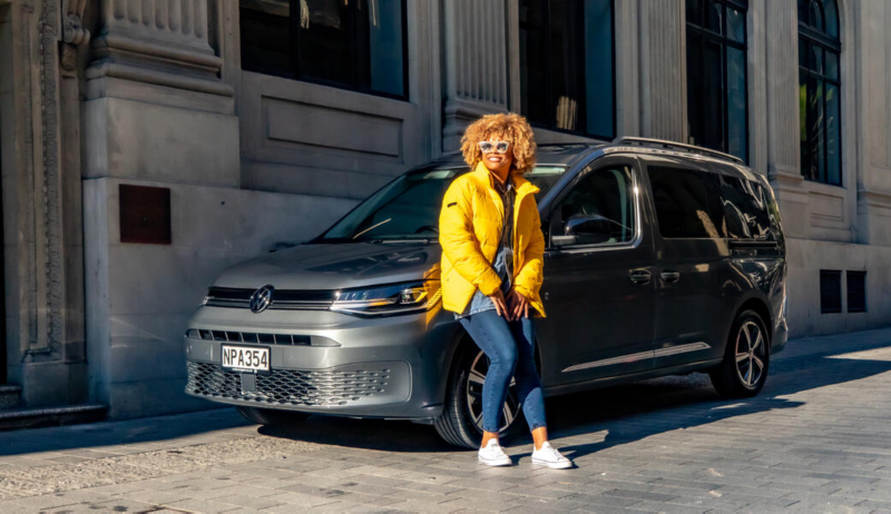 Una mujer apoyada en un VW Caddy estacionado en la calle.