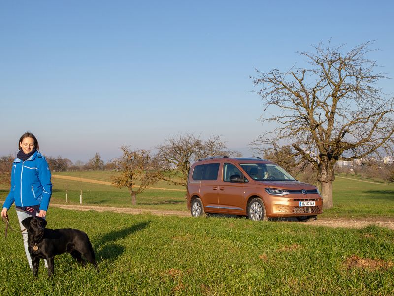 Frau mit Hund steht im Feld vor dem neuen Caddy