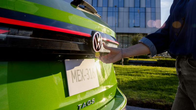 Mano sobre el botón de apertura de la cajuela de un Volkswagen Taos verde, junto al emblema y la placa.