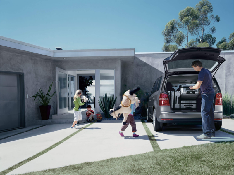 Une famille range des valises dans le coffre d'un VW Touran garé de dos, devant un pavillon.